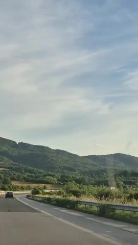 The video captures a scenic drive through a rural landscape. The scene is dominated by rolling green hills covered in lush vegetation, interspersed with trees. A paved road curves through the foreground, with a guardrail separating it from the scenery. A single dark-colored car is visible on the road, moving away from the viewer.

The sky is partly cloudy, with streaks of white clouds against a light blue expanse. The sunlight suggests it is either morning or late afternoon, casting a gentle light on the landscape. The overall impression is one of tranquility and natural beauty. The location cues point to Osikovitsa, Bulgaria, with the rolling hills and verdant scenery characteristic of such an area. There is no visible text in the video.