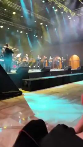 The video shows a band performing on a stage at a live event. The band consists of approximately ten members dressed in black suits. They are playing various instruments, including trumpets, trombones, and a saxophone. The stage is illuminated with bright spotlights and stage lighting creating an atmospheric effect. The audience is visible in the foreground with one person in focus wearing black pants with a pink stripe, seemingly enjoying the performance. The event appears to be held in a large hall, with a patterned floor and a stage set designed with a backdrop mimicking an outdoor architectural style. The camera is hand-held, and there is slight movement throughout the video.