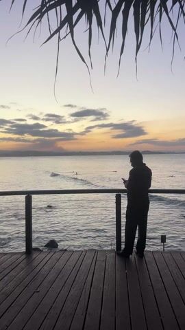 Here's a description of the video: 

The video shows a man standing on a wooden deck overlooking a calm ocean. The man is silhouetted, wearing dark clothing and glasses. He appears to be looking at a phone or other device. The deck has a wooden railing. In the background, the ocean is visible, with several surfers visible on the waves. The sky is a mix of orange, yellow and gray hues, suggesting either sunrise or sunset.  Palm fronds hang down from above, partially obscuring the view. The scene has a peaceful, contemplative atmosphere.