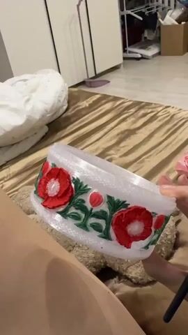 A person's legs are visible on a golden brown textured surface, likely a bed or sofa. They are holding a white cylindrical object decorated with red poppies and green leaves. The person is also holding a black-handled paintbrush in their right hand. In the background, a white dresser or wardrobe and some other household items are visible. The overall environment appears to be indoors.