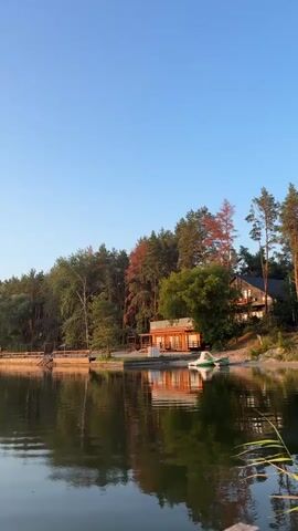 The video shows a calm body of water reflecting a clear blue sky. The shoreline is lined with trees and some structures, including what appears to be a small wooden house and a dock. A white and green boat is visible near the shore. The camera pans slowly across the water, offering a serene and natural landscape. There are no people or animals visible in the video. The time stamp indicates the video is very short, likely a few seconds long.