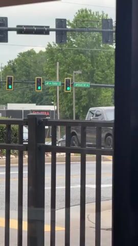 A person films out a window at an intersection where traffic lights display green for both directions. A gray sedan drives through the intersection from left to right. A black metal fence is partially in view in the foreground. Street signs indicate that one direction is named Parker Ct and the other is Davis Rd. The scene is overcast and tree-filled. A building is visible in the background.