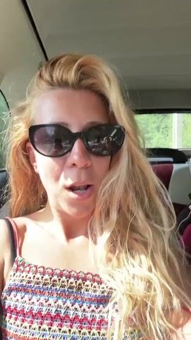 A woman with blonde hair and sunglasses is sitting in a car, talking. She is wearing a colorful strappy top. The background shows the interior of a car and trees visible through the window.