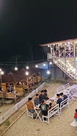 The video shows an outdoor cafe at night. There are several tables and benches set up with people sitting at them. There is a two-story building in the background with lights and people on both levels. String lights are hung around the cafe, illuminating the area. There are also several motorcycles parked along the side of the road. The ground is covered in gravel. Some people are wearing traditional clothing.