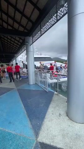 The video shows people boarding a white and red boat at a dock under a covered structure. The dock is painted with blue and grey geometric patterns. The sky is overcast. There are silver pillars supporting the roof of the covered area. The boat is at the edge of the dock, and people are walking onto it. Some people are wearing red life vests. Beyond the boat, the ocean stretches out to a distant shoreline. The camera then moves to show the open ocean. Strong wind noise can be heard.