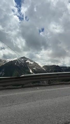 This video shows a scenic mountain landscape with a road in the foreground. The sky is partly cloudy with patches of blue sky visible. Snow-capped mountains rise in the background, with green hills and valleys below. A silver guardrail runs along the side of the road, separating it from a steep drop.

In the second and third segments, a white van with a black cargo carrier on its roof is seen driving past on the road. In the fourth segment, a man is standing on the side of the road, near the guardrail, appearing to be on his phone. He is wearing a t-shirt and pants.

The overall impression is of a journey through a beautiful and mountainous region, possibly a road trip or travel experience. The lighting suggests it is daytime, and the clouds indicate variable weather conditions. The presence of snow on the mountain peaks hints at a location at a higher altitude, and the landscape is consistent with the Gudauri region of Georgia, known for its mountain scenery and ski resorts. There is no visible text in the video.
