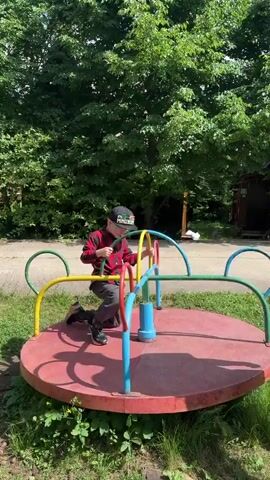 A young boy wearing a red and black plaid shirt, grey pants, a black baseball cap, and glasses is on a red merry-go-round. He is holding onto the yellow and blue bars and appears to be in motion. The merry-go-round is on a dirt path surrounded by grass and lush green trees. The boy's back is mostly to the camera at the beginning of the clip, and he then turns to face more towards the viewer, smiling. The lighting suggests it is daytime.
