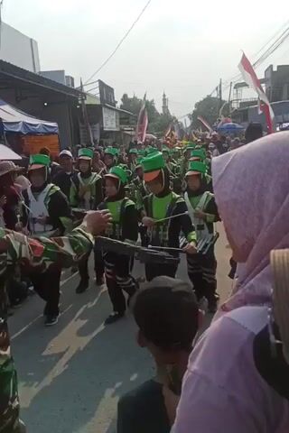 A group of young people in matching green and black uniforms with green hats are marching in a parade. They are playing musical instruments, including what appear to be xylophones. The parade is taking place on a street lined with buildings and stalls. People are watching the parade from the side of the street. Indonesian flags are visible.