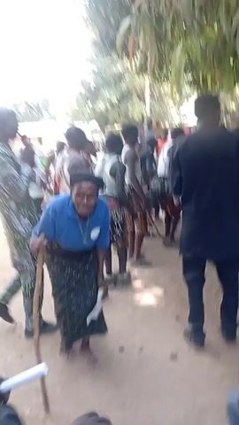 The video shows a gathering of people in an outdoor area with trees. A group of men in traditional attire are dancing to music, some wearing leopard print shorts and vests with white tops.  An older woman in a blue shirt and a patterned skirt stands in the foreground with a cane and a piece of paper.  Other people are standing around watching or sitting in chairs, some wearing traditional hats and clothing.  The ground appears to be dirt.  The overall impression is a community event or celebration.