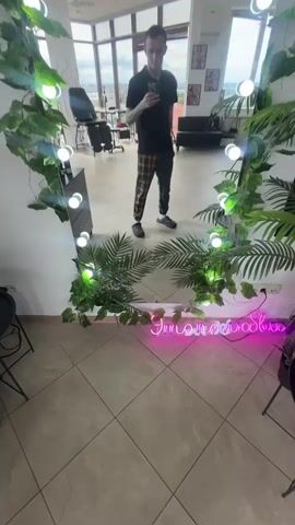 A man stands in front of a full-length mirror, taking a selfie. He is wearing a black t-shirt, checkered pants, and black sneakers. The mirror is decorated with artificial green vines and small white lights. In the reflection, we can see the interior of a room, possibly a studio, with a tattoo chair, a couch, and some artwork on the walls. A pink neon sign that reads "In Love" is visible at the bottom of the mirror. The man makes various hand gestures and facial expressions as he takes the photos.