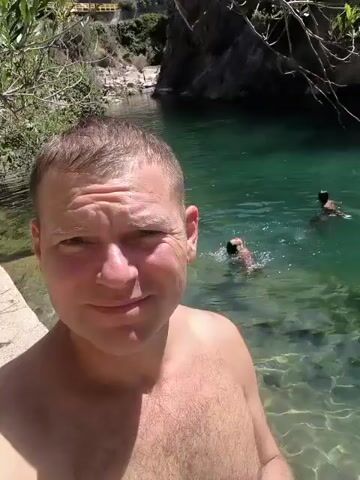A man is taking a selfie video in a river. He is shirtless and smiling at the camera. In the background, the water is clear and blue, and there are rocks and vegetation. Another person is swimming in the river in the distance. The man appears to be enjoying himself.