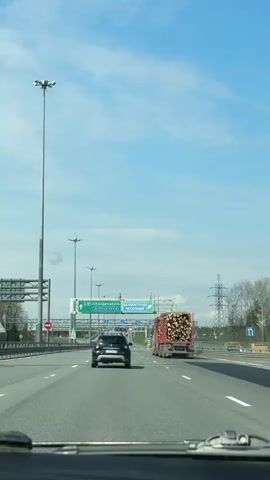 The video shows a view from inside a vehicle traveling on a highway. The sky is bright blue with scattered clouds. In front of the camera are two other vehicles: a black car and a red logging truck loaded with logs. Above the lanes, there are large green signs indicating directions to "SCANDINAVIA/ZELENOGORSK" and "BELOOSTROV/PESOCHNY." Tall light poles stand along the side of the road. The environment appears to be a forested area with power lines visible on the right side of the highway. The vehicle filming the scene is steadily moving forward, maintaining distance from the other vehicles.