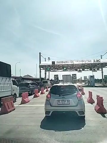 The video shows a car stopped at a toll booth. The toll booth is labeled "GERBANG TOL PADANG TIJI". There are several other cars also waiting to pass through the toll booth. The weather is sunny with a partly cloudy sky. The car in the foreground has the license plate "BL 1719 JO". There are orange plastic barriers on either side of the car. A white truck is visible to the left. There is a building structure visible on the left side of the toll gate. The green arrow lights above the toll lanes are illuminated.