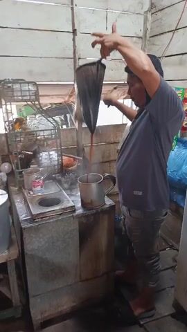 A man stands in a rustic wooden structure, preparing a beverage. He is pouring a dark liquid from a cloth filter, held aloft by his right hand, into a metal pot situated on a stove. Another metal pot is placed next to it. The setting appears to be a small shop or food stall, with shelves holding various packaged goods, including what look like snack items or instant beverages. Steam rises from the stove, indicating heat is being applied. The man is wearing a grey t-shirt and patterned trousers. His posture suggests he is actively engaged in the pouring process. The overall environment is simple and functional, likely indicative of a local eatery.