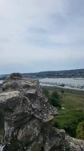 The video shows a scenic view from a stone structure, possibly a castle. The camera first pans across the landscape, revealing a river or bay dotted with numerous boats and a town in the distance. The foreground consists of rocky formations and some greenery. The camera then moves along a stone pathway, lined with a white metal railing, leading towards an archway in a tall stone wall. A sign is partially visible at the end of the railing. The sky is partly cloudy.