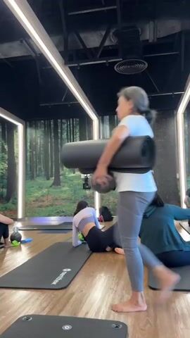 The media shows a group of people participating in a yoga class in an indoor studio. The studio has a wooden floor and is decorated with a large mural of a forest on the back wall. There are several yoga mats laid out on the floor, and some participants are already in various poses. One person is walking into the frame carrying two rolled-up yoga mats. Another person is in a kneeling position, while two others are seated on their mats. The lighting appears to be artificial, with bright, strip lights illuminating the space. The overall atmosphere seems calm and focused, typical of a yoga or fitness class. The time of day is not specified, but the indoor setting suggests it could be any time. The location is identified as Nong Pa Khrang, Thailand. There are no visible weather conditions. One visible text on a yoga mat says "Wamiyoga".