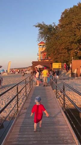 A child in a red shirt runs down a wooden pier towards a beach. People are visible on the sandy beach in the background, some are sitting on chairs, others are walking around. There are trees and a building with a red roof on the right side of the image. The sun is setting, casting a warm glow on the scene.