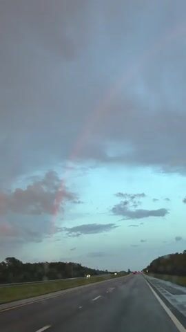 A video captures a view from a vehicle driving on a wet highway. The sky is filled with gray and blue clouds, with a faint pink rainbow arching across the upper portion of the frame. The highway is empty except for a few distant cars and a black car in the left lane. The trees on the right side of the road are dark and indistinct. The video appears to be taken during dusk or after a rain shower.