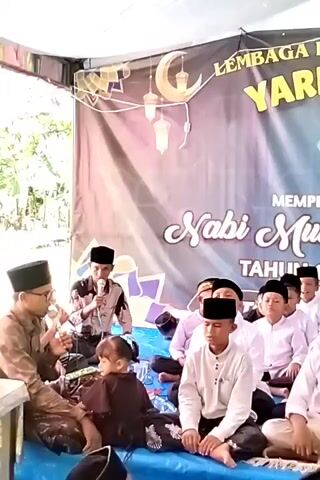 The video shows a group of boys and two men in an outdoor setting, presumably a religious gathering. The boys, dressed in white shirts and black caps, are sitting on a blue mat on the ground. Two men are also present, one holding a microphone and appearing to be speaking, while the other is sitting near him. Behind them is a banner with the words "Lembaga Pendidikan Islam YARHAMUNA," "Memperingati Maulid Nabi Muhammad SAW," and the date "Tahun 1447 H/ 2025 M." The banner also features a crescent moon and star symbol, as well as a logo with Arabic script.  The setting suggests a religious ceremony commemorating the birth of the Prophet Muhammad. A young girl sits among the boys.