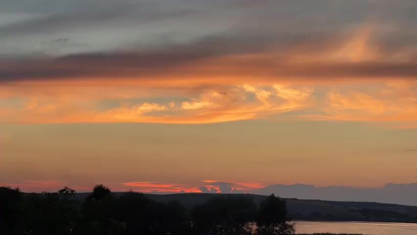 The video shows a sunset over a body of water, possibly a river or lake, with a silhouetted landscape in the foreground. The sky is filled with dramatic clouds, painted in shades of orange, pink, and grey, reflecting the warm hues of the setting sun. The water's surface appears calm, with subtle ripples, and the distant shore is dotted with a few trees and possibly some structures, creating a picturesque and serene scene. The overall atmosphere is peaceful and tranquil.