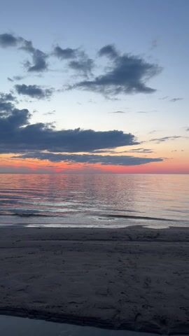The video shows a serene beach scene at dusk. The sky transitions from blue at the top to a soft pink and orange hue near the horizon, reflected in the calm water. Dark, grey clouds are scattered across the sky. The beach is sandy and still, with gentle waves lapping at the shore. The scene appears tranquil and undisturbed.