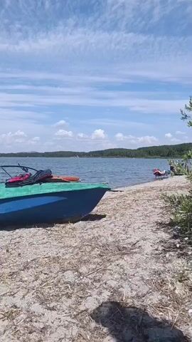 A woman in a bikini and hat walks away from the camera on a sandy beach towards a lake. A blue and green boat is parked on the beach to her left, with bags inside. The lake is calm, with trees and hills in the background. The sky is blue with some white clouds.