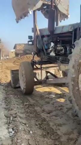 The video shows a blue tractor moving along a dirt road or path. The tractor has large, muddy tires. It also has a metal blade or leveler attached to the front and a mechanical arm with a bucket on top. The tractor appears to be working on the road, possibly leveling it or clearing dirt. The background shows a clear blue sky and a few buildings in the distance.