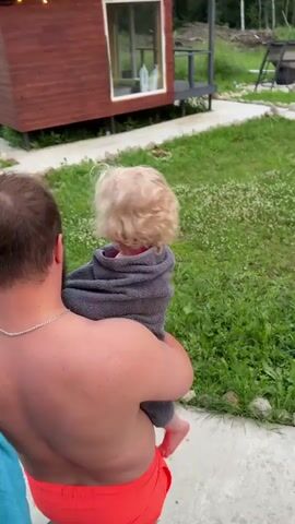 A man in red swim trunks and a blue towel draped over his shoulder carries a blonde-haired child in a gray towel. They are walking on a concrete path bordered by grass and stones. A small wooden structure with windows is visible in the background, along with trees and fencing. The scene appears to be outdoors in a backyard setting.