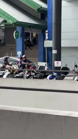 The video shows a group of people riding motorcycles. The motorcycles are parked side-by-side and some are in motion. The people are wearing helmets. The environment appears to be an urban area with a building in the background. There are also signs visible, one of which indicates no parking.