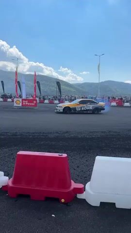A drift race event is taking place. A white, yellow, and black BMW drift car with the number 37 and "ROWE" branding is shown drifting, emitting blue smoke from its tires. Spectators are visible in the background, as are mountains and a bright blue sky with clouds. Red and white plastic barriers line the track in the foreground.