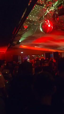 The video shows a crowded indoor party. The room is dimly lit, with colorful lights in red, green and blue. There is a disco ball hanging from the ceiling. The camera angle is looking up at the ceiling and across the crowd of people. The music playing appears to be dance music.