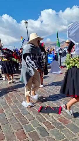 A group of people in traditional clothing are dancing in a cobblestone square. The sky is blue with scattered clouds. In the foreground, a person in a wide-brimmed hat and a checkered shirt with a grey cape dances on small wheeled shoes. Behind them, other dancers in colorful costumes move with white flags and greenery. In the background, onlookers are visible behind a barrier, and flags are flying. The scene appears to be a festive outdoor event or parade.