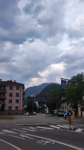 The video shows a cloudy sky over a town with mountains in the background. A street intersection is visible in the foreground with a pink building on the left and a white building on the right. There are traffic signs, a pedestrian crossing, and a bicycle lane marked on the road. A few vehicles are present, including a motorcycle and a truck. Trees line the street, and the overall atmosphere appears to be overcast.
