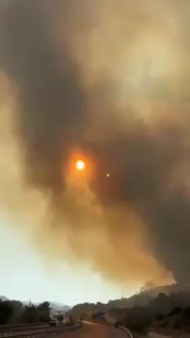 The video shows a large forest fire emitting plumes of smoke that darken the sky. The sun is visible through the smoke, appearing as a bright orange disc.  The fire is burning on a hillside covered with trees and vegetation, with flames visible in some areas.  A highway runs along the base of the hill, and vehicles, including tractor trailers, are driving on the road. A few people stand along the edge of the road, observing the fire.