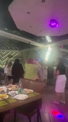 This media file captures a lively scene inside a restaurant or club in Orsha, Belarus, likely during an evening event. The primary subjects are a group of people dancing and socializing, illuminated by dynamic, colorful disco lights that strobe across the room. 

In the foreground, a table is set with plates of food and drinks, suggesting a dining or celebratory occasion. The table is draped with a green tablecloth. Behind it, patrons are actively engaged in dancing. A woman in a bright green shirt is particularly prominent, with her back to the camera as she dances. Other individuals are seen in various states of movement, some interacting with each other.

The setting appears to be a modern establishment with a distinctive ceiling featuring geometric patterns and recessed lighting. A bar area with bottles of alcohol is visible in the background, further indicating a place for drinks and entertainment. The overall atmosphere is energetic and festive, with the colorful lights contributing to a party-like mood. While no specific time of day is explicitly stated, the lighting and activity suggest it is nighttime, consistent with typical evening entertainment venues. The weather is not discernible from the indoor scene. No legible text is visible in the frame.