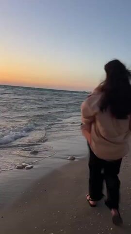 A person with long dark hair, wearing a light brown jacket, black pants, and dark sandals, walks along a sandy beach at dusk. The person initially walks away from the camera, parallel to the water's edge with small waves breaking. They then turn to their left, facing the camera and the ocean, and adjust their hair or clothing before standing still, looking forward. The sky displays sunset colors of orange, pink, and light blue, with distant lights visible on the horizon.