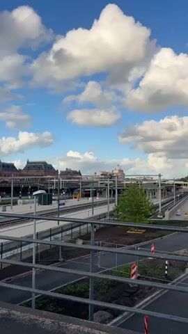The video pans from a train station with multiple tracks and platforms to a modern office building. The sky is blue with scattered white clouds. The train station appears to be under construction or renovation, with yellow barriers and construction materials visible on some platforms. A red and white no parking sign is visible on a pole in the foreground. The office building is tall and rectangular with many windows. The camera then pans further to the right, showing more of the train station and surrounding buildings.