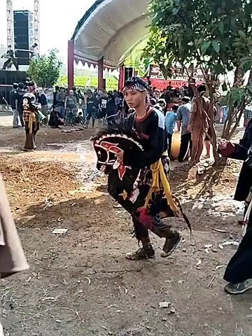 The video shows an outdoor event. In the foreground, a person dressed in traditional clothing, possibly a performer, is holding a prop that resembles a bull's head and body. They are wearing a bandana, a black shirt with white and yellow accents, and patterned pants. The performer is facing towards the right of the frame. Behind them, a crowd of people is gathered. In the background, there is a large speaker system mounted on a truss structure. Several people are visible near the speakers, and some are standing in front of a tent-like structure with a white roof and red trim. There are also flags visible, including one that appears to be the Indonesian flag. The ground is dirt and appears somewhat muddy in places. The overall atmosphere suggests a cultural performance or festival.