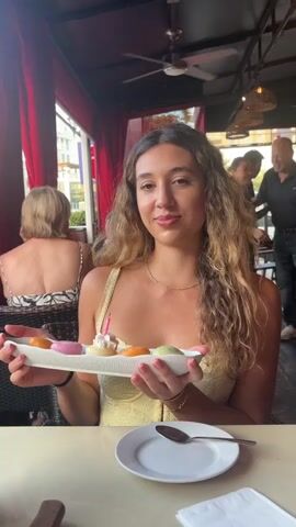 A young woman with long curly blonde hair, wearing a yellow top, is holding a white oval dish with five small pastel-colored desserts and a pink birthday candle. She is seated at a table in what appears to be an outdoor restaurant. A white plate with a spoon rests on the table in front of her. In the background, several other people are visible, and red curtains hang behind the woman. The lighting suggests it is daytime.