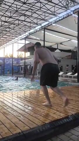A person is captured in mid-air jumping into a pool, creating a large splash. The person is wearing black shorts and is facing away from the camera. The jumping platform is made of wet wooden planks. The pool appears to be an indoor or covered outdoor swimming area with a metal roof structure. The background shows a blue advertisement banner, people swimming in the pool, and lounge chairs. The lighting suggests it is either early morning or late afternoon with the sun visible in the distance.