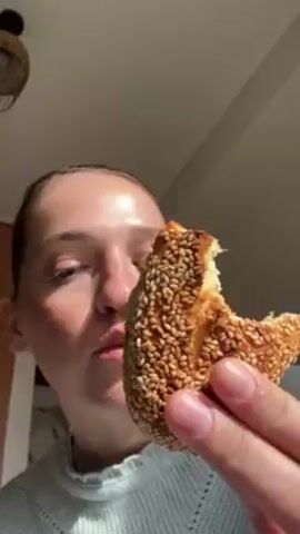 The video shows a woman eating a sesame-covered pastry, presumably a bagel or bun, filled with some sort of soft filling. The camera is close-up, focusing on her face and hand as she takes a bite. The pastry is golden brown and covered in sesame seeds, with a white filling visible. The woman is fair-skinned, with her hair pulled back. She's wearing a light blue knitted sweater. Her hand has a ring on one of the fingers. The background is slightly blurred, but suggests an interior setting, possibly a kitchen, with a light fixture visible in the top left corner. The lighting appears to be bright, possibly from natural sunlight.