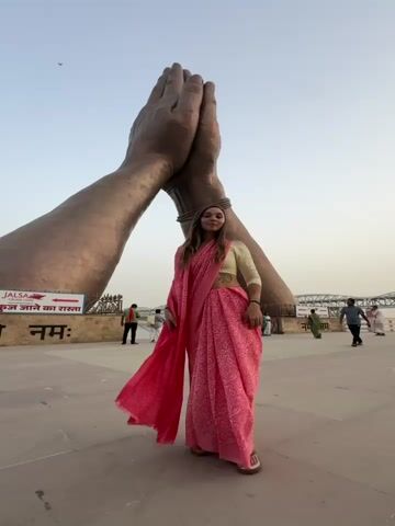A woman in a pink sari stands in front of a large, bronze-colored sculpture of two hands pressed together in a prayer position. She raises her arms above her head, as if reaching for the sculpture. The background shows a wide open space with other people walking around, and a river in the distance. A sign in Hindi is visible near the hands. The sky is a clear, pale blue.
