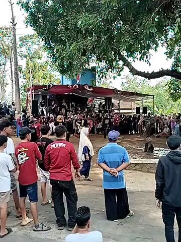 The video shows an outdoor performance with a stage set up under a large tree. A crowd of people is gathered, watching the performance. On the stage, several individuals are dressed in traditional costumes, appearing to be performers. Some are holding instruments, suggesting a musical or dance performance. The stage is decorated with red drapes and banners, one of which has a logo with the text "GUNTI" visible. People in the foreground are observing the event. The environment appears to be a rural or semi-rural area, with vegetation and simple structures in the background. The lighting suggests it is daytime.
