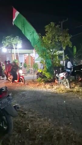 A group of people are gathered outside at night. There are several motorcycles parked. A large green flag with red and white at the top flies above the scene. A person is riding a motorcycle with its headlights on, moving through the gathering. The area is lit by artificial lights.