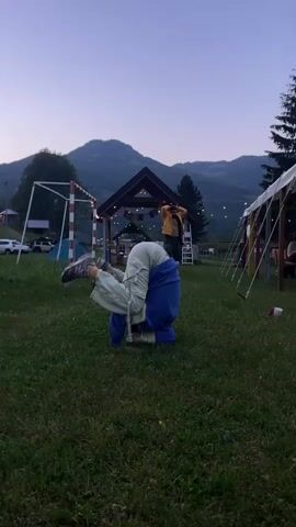Here is a description of the video:
A person in blue and green clothing is performing a headstand on a grassy lawn. In the background, there is a structure with lights, a soccer goal, a tent, trees and mountains. A person in a yellow jacket is visible in the background, standing and observing.  The sky is clear and bright, suggesting it is daytime.