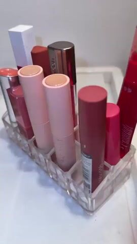 A close-up shot showcases a clear acrylic organizer filled with various lipsticks and lip glosses. The lipsticks are arranged in a row, with tubes of different colors and sizes. Some of the lipsticks are pink, red, and coral. The organizer is placed on a white surface, possibly a countertop or vanity. In the background, a glass vase containing artificial white flowers and greenery is visible. The lighting is soft, creating a warm and inviting atmosphere. The camera slowly pans across the lipsticks, highlighting their packaging and colors. The video ends with a focus on the different shades of red and pink lipsticks in the organizer.