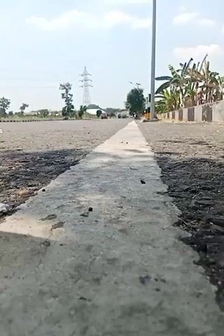 This is a first-person perspective video of an asphalt road with a white line painted down the center. The camera is low to the ground, close to the white line, and appears to be moving forward. The road is somewhat worn and there are some debris on the left side. In the background, there are trees, a power line tower, some buildings, and banana trees. The sky is clear and blue with some clouds. There are a few cars visible in the distance, as well as a street light on the right side of the road.