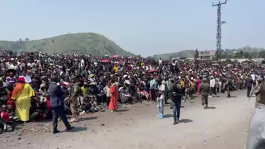 A wide shot shows a large crowd of people gathered in an open area, with a grassy hill visible in the background. Most of the crowd is seated, with individuals standing and walking amongst them. Several men in suits and uniformed military personnel are also present, seemingly providing security or direction. The ground appears to be dusty or gravelly. The sky is clear and bright, suggesting a sunny day. A tall communication tower stands in the distance. The atmosphere seems to be charged with anticipation or excitement, although the specific event or purpose of the gathering is unclear from the visual alone.
