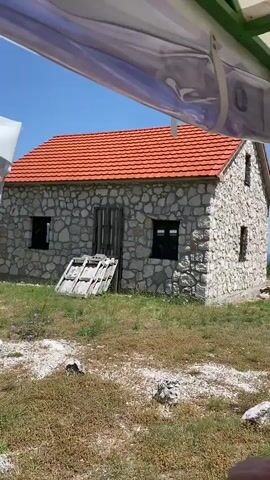 The video pans across a rustic stone building with a red tiled roof. The building has two windows on the front facade and one on the side. There is a wooden door on the front. To the left of the door, there are some wooden planks stacked against the wall. The building is situated on a grassy hill with some rocks scattered around. A small tree grows in front of the building, and further to the right, a large leafy tree obscures some of the view. In the background, rolling hills and a bright blue sky with some clouds are visible. The video appears to be taken from under a white canopy or awning.