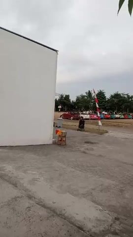 A brief video shows an outdoor scene with a white building on the left. In the middle ground, a person is standing next to a tall pole with an Indonesian flag. A row of colorful cars is parked in the background, and trees are visible behind the cars. The ground in the foreground appears to be concrete or pavement. The sky is overcast.